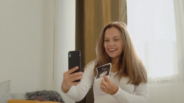 Donna incinta mostra all'interlocutore una foto dall'esame ecografico del feto utilizzando un collegamento video su uno smartphone — Video Stock