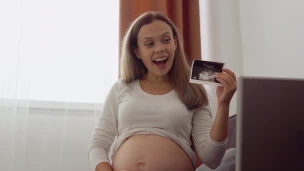 Terhes, világos bőrű nő megmutatja a beszélgetőpartner egy fényképet az ultrahang vizsgálata a magzat segítségével videokommunikáció egy laptop — Stock videók