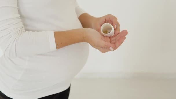 Mulher grávida de pele clara fica em um fundo branco e derrama pílulas na palma da mão — Vídeo de Stock