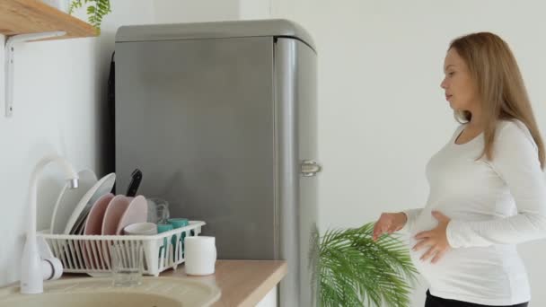 Беременная женщина подходит к холодильнику, берет миску нарезанных овощей и улыбается в камеру — стоковое видео
