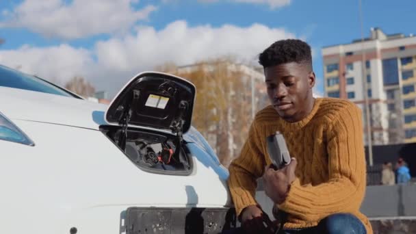 Le conducteur mâle afro-américain connecte la voiture électrique au système d'alimentation pour charger la batterie de la voiture — Video