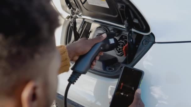 Африканський американець чоловічий водій з'єднує електричний автомобіль з системою живлення, щоб зарядити акумулятор автомобіля і контролювати процес зарядки через свій смартфон. — стокове відео