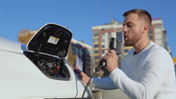 En ljushyad manlig förare håller en laddkabel för en elbil som sitter nära ett öppet bilbatteri och blåser ut inbillad rök från laddkabeln, som efter ett skott — Stockvideo