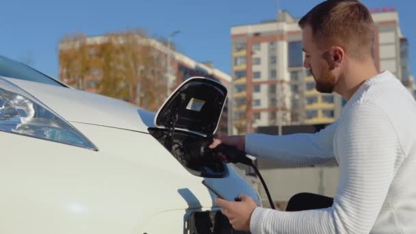 건강 한 남자 운전자가 전기 자동차를 전력 시스템에 연결하여 자동차 배터리를 충전하고 스마트폰을 통해 충전 과정을 제어 한다 — 비디오