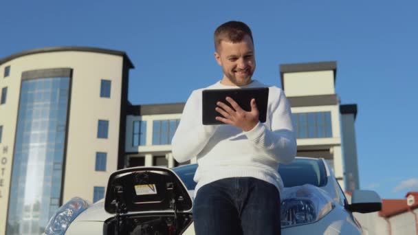 Een man met een lichte huid staat met een tablet in zijn handen naast een elektrische auto terwijl hij oplaadt — Stockvideo