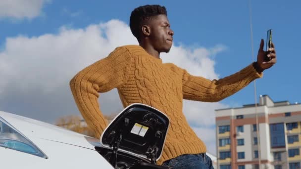 En svart förare fotograferas på en smartphone kamera står nära en vit elbil — Stockvideo