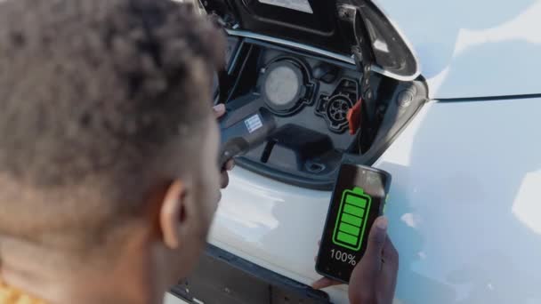 Un conductor masculino de piel oscura conecta un coche eléctrico al sistema de energía para cargar la batería del coche y controla el proceso de carga a través de su teléfono inteligente — Vídeo de stock