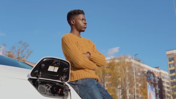Темношкірий водій стоїть біля білого електромобіля під час заряджання батареї — стокове відео