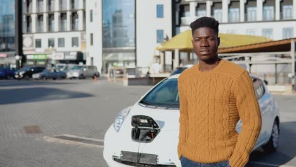 Αφροαμερικάνος οδηγός στέκεται κοντά σε ένα λευκό ηλεκτρικό αυτοκίνητο, ενώ η φόρτιση της μπαταρίας — Αρχείο Βίντεο
