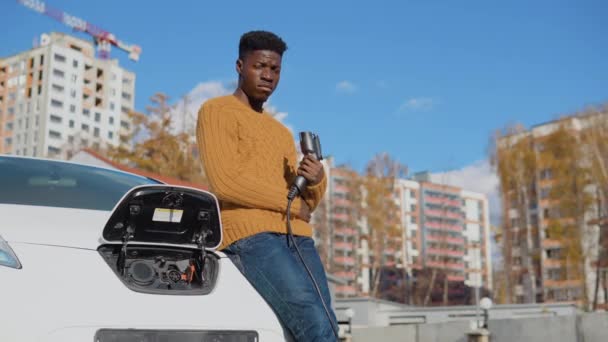 Un șofer de sex masculin cu piele închisă deține un cablu de încărcare pentru o mașină electrică care stă lângă bateria deschisă a unei mașini — Videoclip de stoc