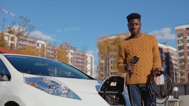 Conductor masculino afroamericano sostiene un cable de carga para un coche eléctrico de pie cerca de la batería abierta de un coche — Vídeos de Stock