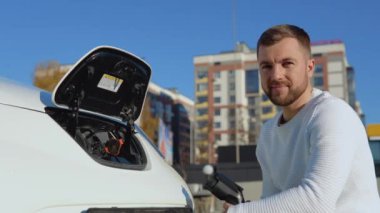 Bir erkek sürücü açık bir araba aküsünün yanında otururken elektrikli bir araba için şarj kablosu tutar ve eliyle iyi bir işaret gösterir.