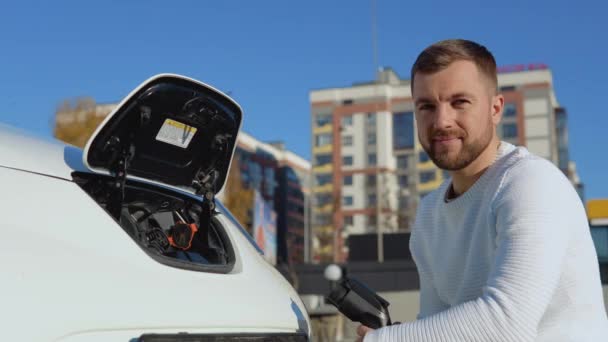 Egy férfi sofőr egy elektromos autó töltőkábelét tartja, miközben nyitott akkumulátor mellett ül, és jó jelet mutat a kezével. — Stock videók