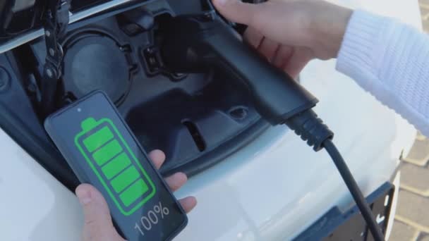 Řidič se světlou pletí připojí elektrický automobil k napájecímu systému, nabíjí baterii a ovládá nabíjecí proces pomocí svého chytrého telefonu. — Stock video