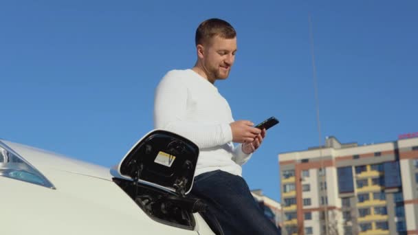 Un conductor masculino conecta un coche eléctrico al sistema de energía para cargar la batería del coche y controla el proceso de carga a través de su teléfono inteligente — Vídeos de Stock