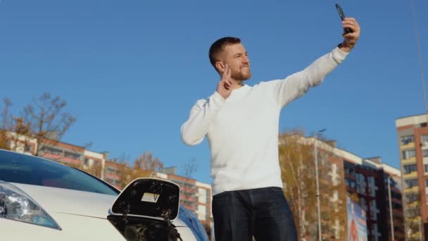 Un conductor es fotografiado en una cámara de teléfono inteligente de pie cerca de un coche eléctrico blanco — Vídeos de Stock