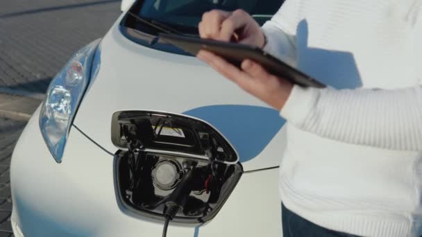 Muž řidič stojí s tabletem v ruce u elektrického auta, zatímco se nabíjí — Stock video