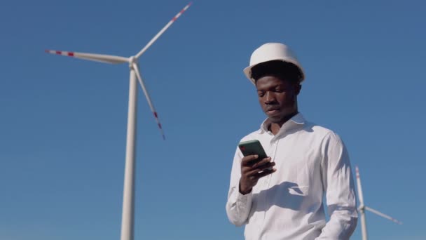 Ingegnere elettrico afroamericano si trova sullo sfondo di un mulino a vento in una centrale elettrica ad aria e ha una conversazione telefonica. turbine eoliche che generano energia rinnovabile pulita — Video Stock