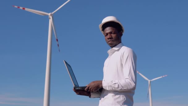 Ingegnere elettricista afroamericano in piedi sullo sfondo di un mulino a vento in una centrale elettrica ad aria con una tavoletta in mano — Video Stock