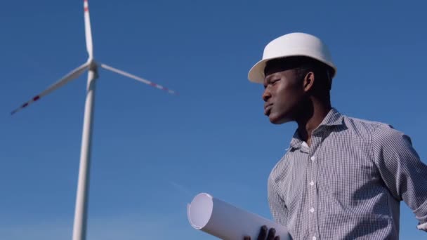 Ingeniero eléctrico afroamericano en un casco y con un dibujo en la mano se encuentra en el fondo de un molino de viento en una planta de energía aérea. Concepto de energía renovable — Vídeo de stock
