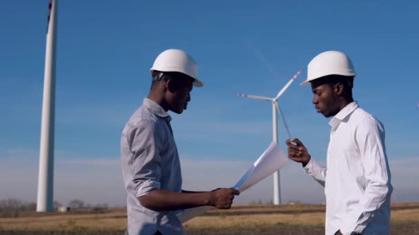 Dva muži afroameričtí elektrotechnici stojí na pozadí větrného mlýna ve vzdušné elektrárně. Drží kresbu v rukou a zkoumají ji. — Stock video