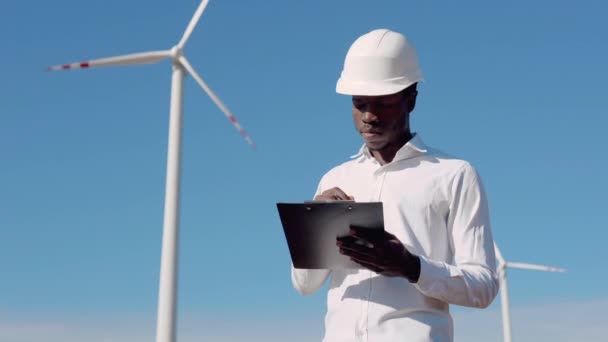 Bir hava santralinin arka planında duran Afrikalı elektrikçi mühendisi kayıtları tutuyor. Rüzgar güç türbinleri temiz yenilenebilir enerji üretiyor — Stok video