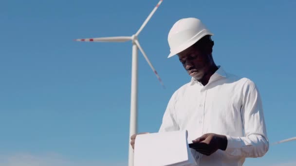 Engenheiro elétrico afro-americano em pé no fundo de um moinho de vento em uma usina de energia aérea registros de leitura — Vídeo de Stock