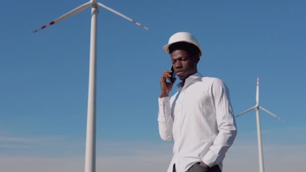 Ingegnere elettrico afroamericano si trova sullo sfondo di un mulino a vento in una centrale elettrica ad aria e ha una conversazione telefonica. Concetto di energia rinnovabile — Video Stock