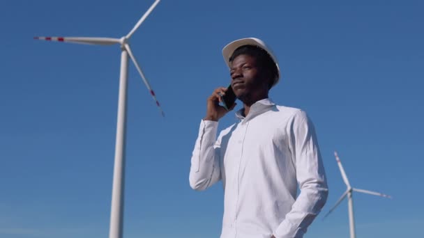 Engenheiro elétrico afro-americano está contra o pano de fundo de um moinho de vento em uma usina de energia aérea e tem uma conversa telefônica — Vídeo de Stock