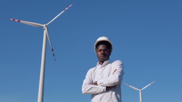 Ingegnere elettricista afroamericano si trova sullo sfondo di un mulino a vento in una centrale elettrica ad aria e mostra un pollice verso l'alto. turbine eoliche che generano energia rinnovabile pulita — Video Stock