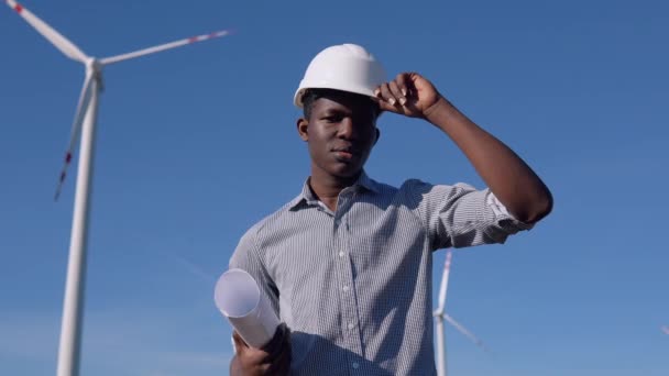 Ingeniero eléctrico afroamericano en un casco y con un dibujo en la mano está parado en el fondo de un molino de viento en una planta de energía aérea — Vídeo de stock