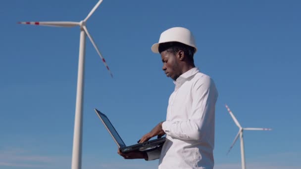 Αφροαμερικάνος ηλεκτρολόγος μηχανικός στέκεται στο φόντο ενός ανεμόμυλου σε ένα εργοστάσιο παραγωγής ηλεκτρικής ενέργειας αέρα με ένα δισκίο στα χέρια του — Αρχείο Βίντεο