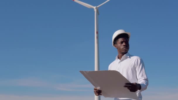 Der afroamerikanische Elektriker mit Helm steht vor der Kulisse einer Windmühle in einem Luftkraftwerk und liest eine Zeichnung. Windkraftanlagen zur Erzeugung sauberer erneuerbarer Energien