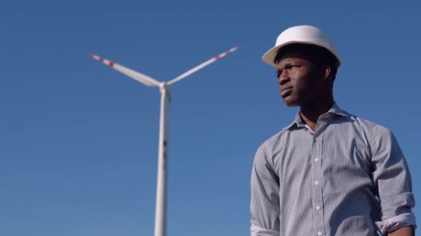 Afroamerický elektrikář v helmě stojí na pozadí větrného mlýna ve větrné elektrárně a dívá se z obzoru na kameru — Stock video