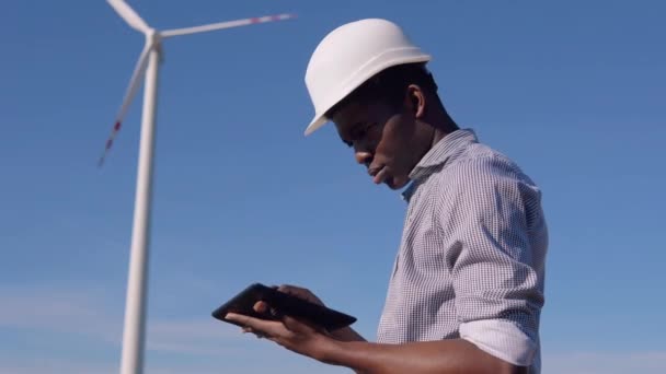 Un electricista afroamericano en un casco se levanta contra el telón de fondo de un molino de viento en una planta de energía aérea y mira una tableta y cruza la pantalla. Concepto de energía renovable — Vídeo de stock
