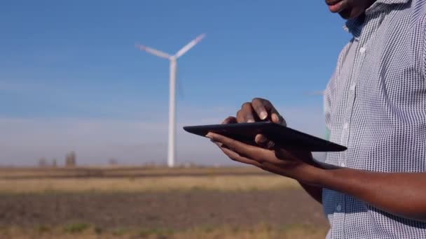 ヘルメットを被ったアフリカ系アメリカ人の電気技師は、風力発電所の風車を背景に立っており、画面全体にタブレットとスワイプを見ています。手とタブレットの近景 — ストック動画