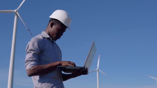 Un ingeniero eléctrico afroamericano está de pie contra el telón de fondo de un molino de viento en una planta de energía aérea con una computadora portátil en sus manos y escribiendo algo — Vídeo de stock