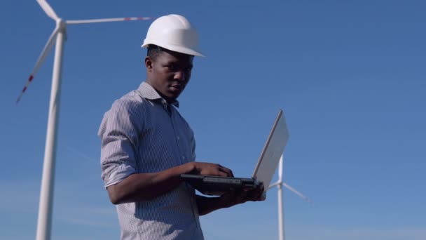 Un ingegnere elettrico afroamericano si trova sullo sfondo di un mulino a vento in una centrale elettrica ad aria con un computer portatile in mano e sta scrivendo qualcosa. Concetto di energia rinnovabile — Video Stock
