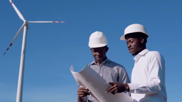 Dois engenheiros elétricos afro-americanos masculinos estão contra o pano de fundo de um moinho de vento em uma usina de energia aérea com um desenho em suas mãos e lê-los — Vídeo de Stock