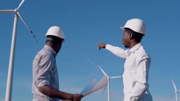Due ingegneri elettrici afroamericani di sesso maschile si trovano sullo sfondo di un mulino a vento in una centrale elettrica ad aria con un disegno in mano e discutono questioni tecniche — Video Stock