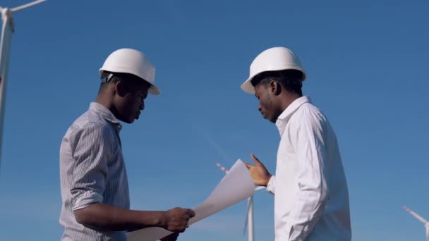 Два афроамериканских инженера-электрика стоят на фоне ветряной мельницы на воздушной электростанции с рисунком в руках и обдумывают технические вопросы. — стоковое видео