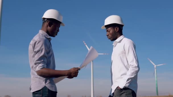 Dos ingenieros eléctricos afroamericanos masculinos se paran contra el telón de fondo de un molino de viento en una planta de energía aérea. Ellos sostienen el dibujo en sus manos y lo examinan — Vídeo de stock
