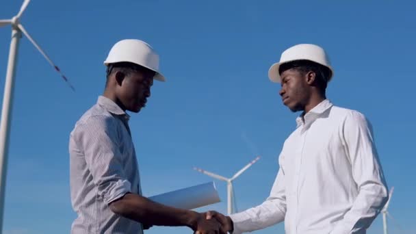 Due ingegneri elettrici afroamericani di sesso maschile stanno sullo sfondo di un mulino a vento in una centrale elettrica ad aria e si stringono la mano — Video Stock