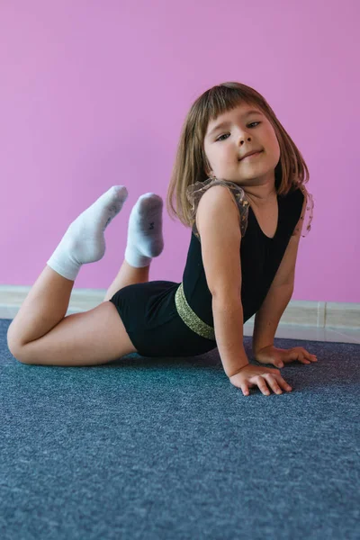 Little girl in black shows exercises for body flexibility on a background of a pink wall. The child is a gymnast