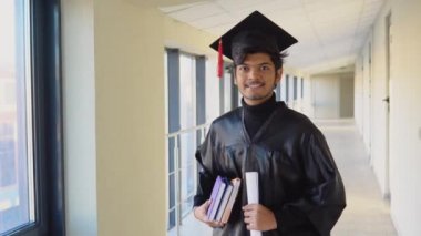 Indian graduate in mantle stands with a diploma and books in her hands and smiles. An important event. Young specialist