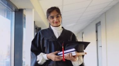Pakistani female graduate in mantle stands with a books in her hands and smiles