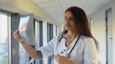 Female indian doctor or intern examines x-ray of lungs, holding it in hands indoors. Specialist holds transparent image of chest in arms, and carefully researches it
