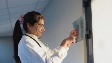Female indian doctor or intern examines x-ray of lungs, holding it in hands indoors. Specialist holds transparent image of chest in arms, and carefully researches it