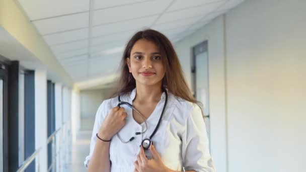 Friendly happy indian woman physician or nurse professional general practitioner posing with stethoscope looking at camera in clinic. Smiling young female doctor close up portrait — 图库视频影像