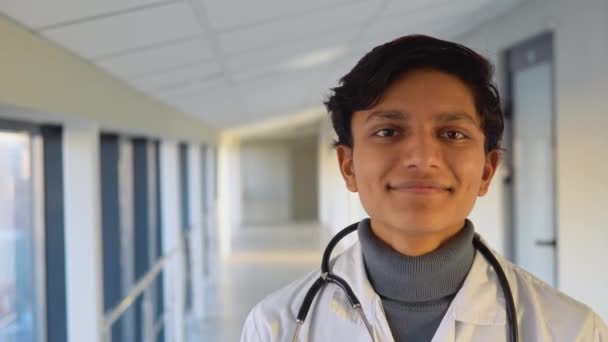 Friendly happy indian male physician professional general practitioner posing with stethoscope looking at camera in clinic. Smiling young doctor close up portrait — 图库视频影像